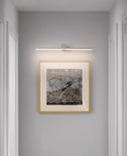 A lit Clérachic wall light glows above a gold-framed abstract print in a hallway.