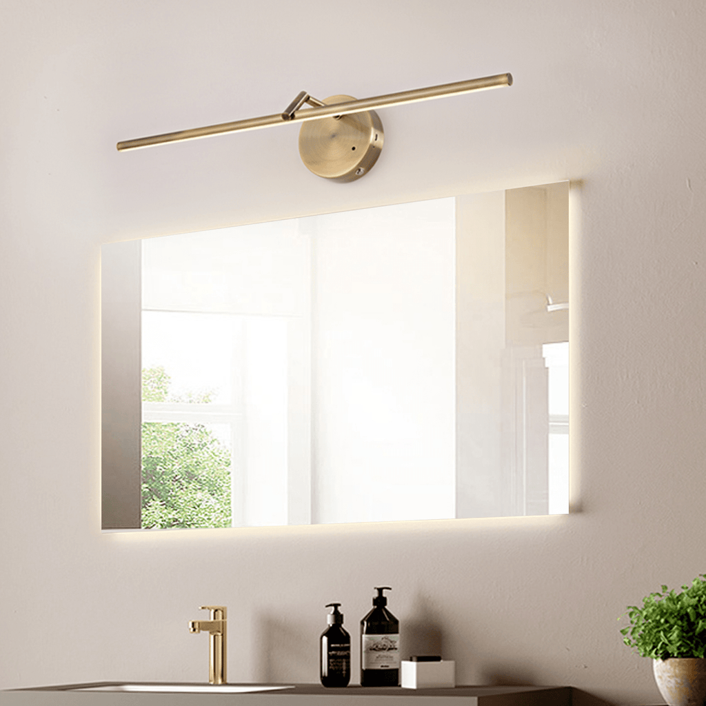 This gold-finish Clérachic wall light is mounted beside a large frameless mirror above a bathroom sink with a brass tap and toiletries.
