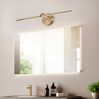 This gold-finish Clérachic wall light is mounted beside a large frameless mirror above a bathroom sink with a brass tap and toiletries.
