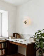 A Clérachic alabaster wall light glows above a marble-topped desk with stacked books, a bookshelf, and a potted plant.