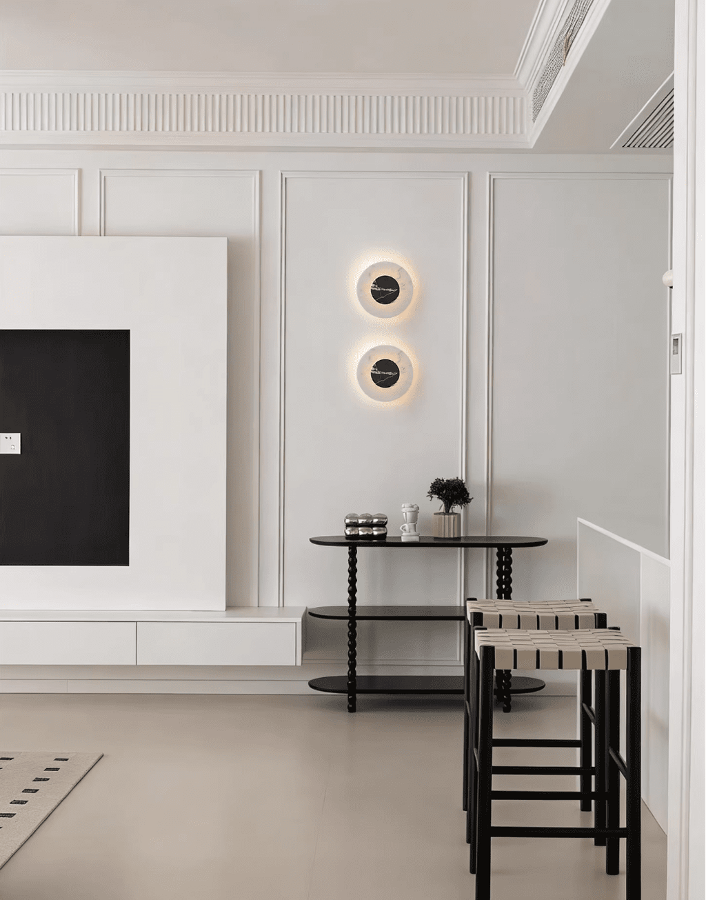 Two Clérachic alabaster wall lights lit on a panelled wall beside a media unit, above a black bar table and stools.