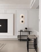 Two Clérachic alabaster wall lights lit on a panelled wall beside a media unit, above a black bar table and stools.