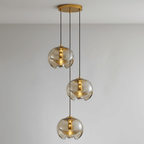Three amber glass shades with gold fittings hang at staggered heights from a round gold ceiling plate, this Clérachic fixture shown against a plain wall.