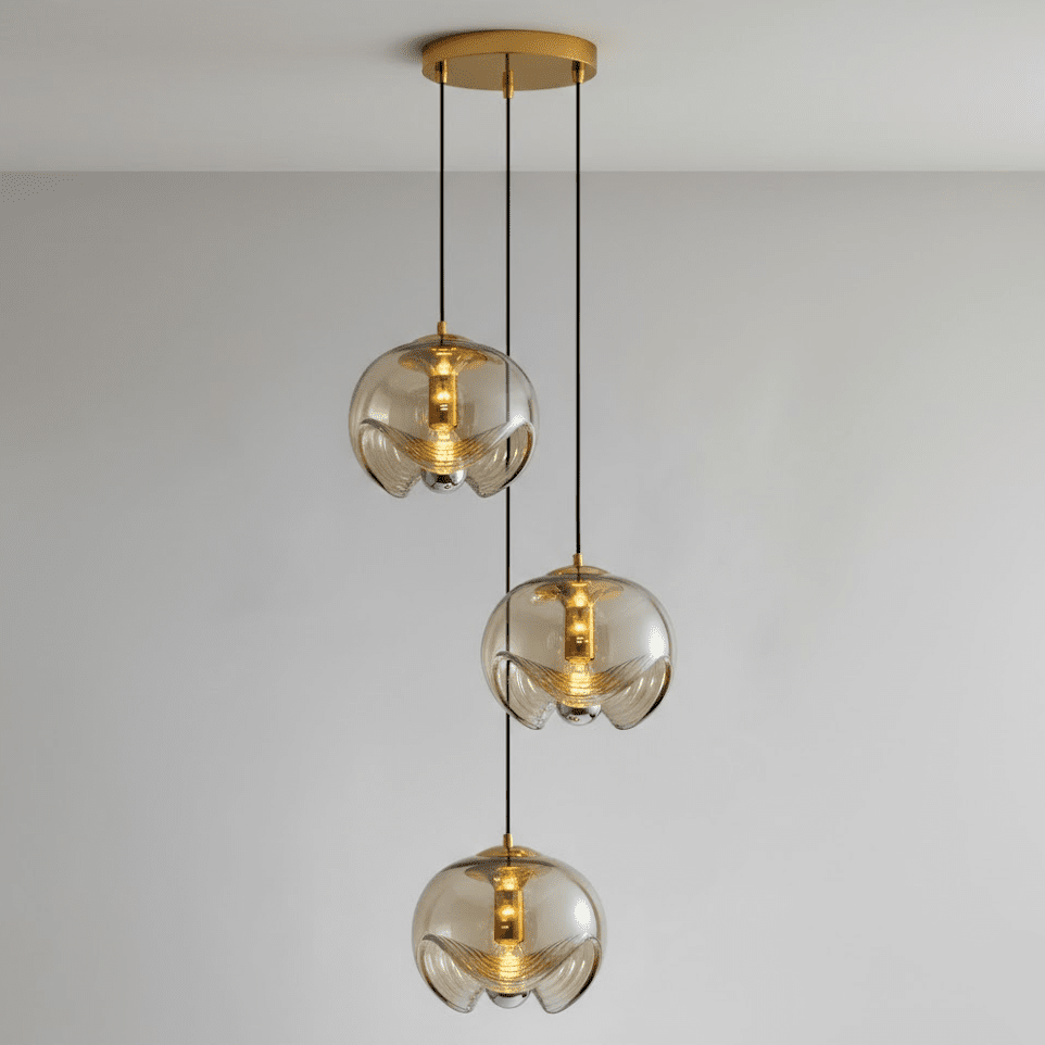 Three amber glass shades with gold fittings hang at staggered heights from a round gold ceiling plate, this Clérachic fixture shown against a plain wall.