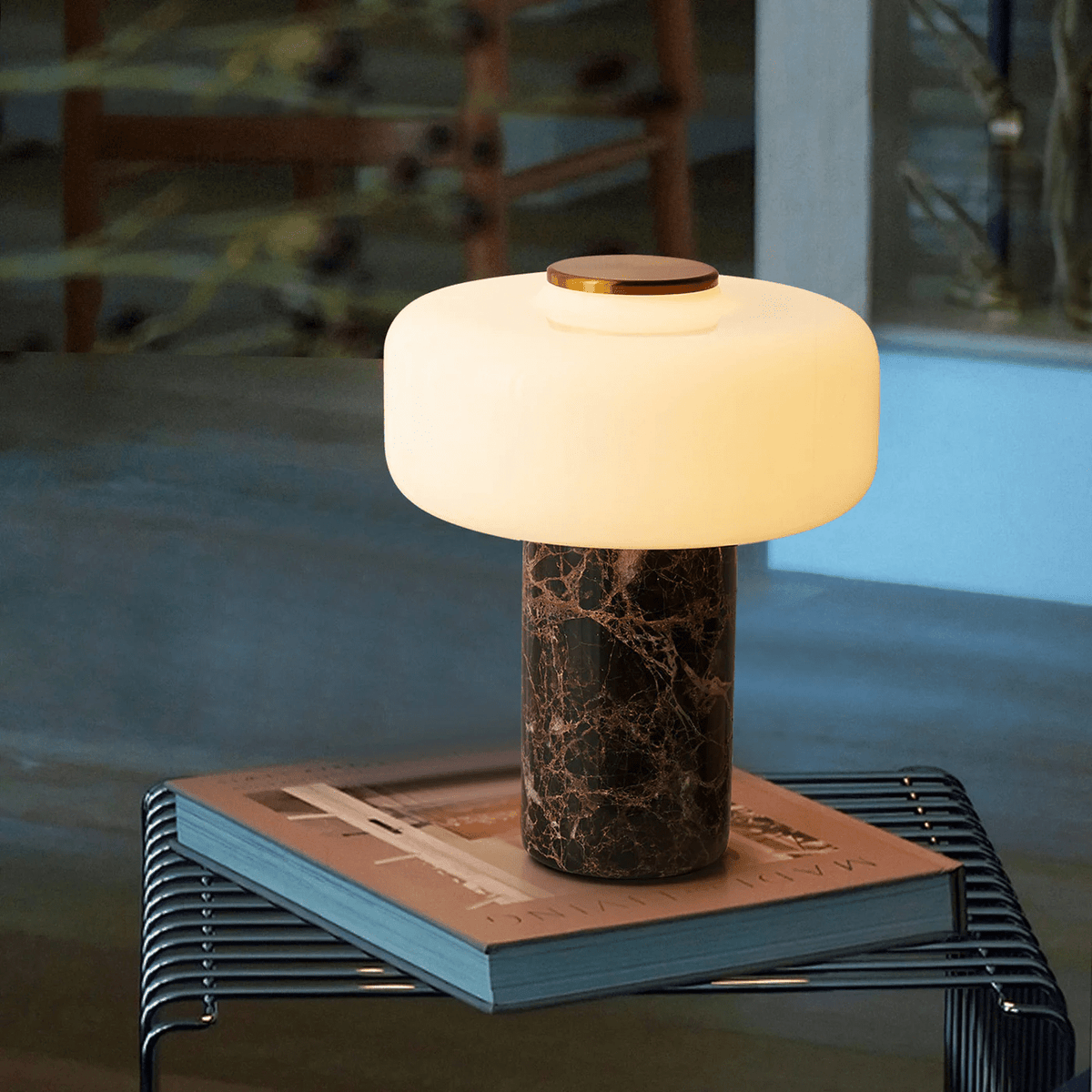 This Clérachic table lamp rests on a stack of books on a wire side table, dusk light through the window behind.