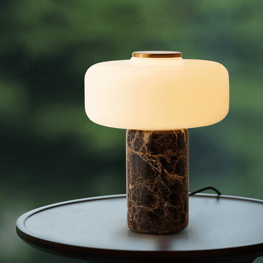 On a round black side table, blurred greenery behind it, a Clérachic table lamp with a dark marble base stands lit.