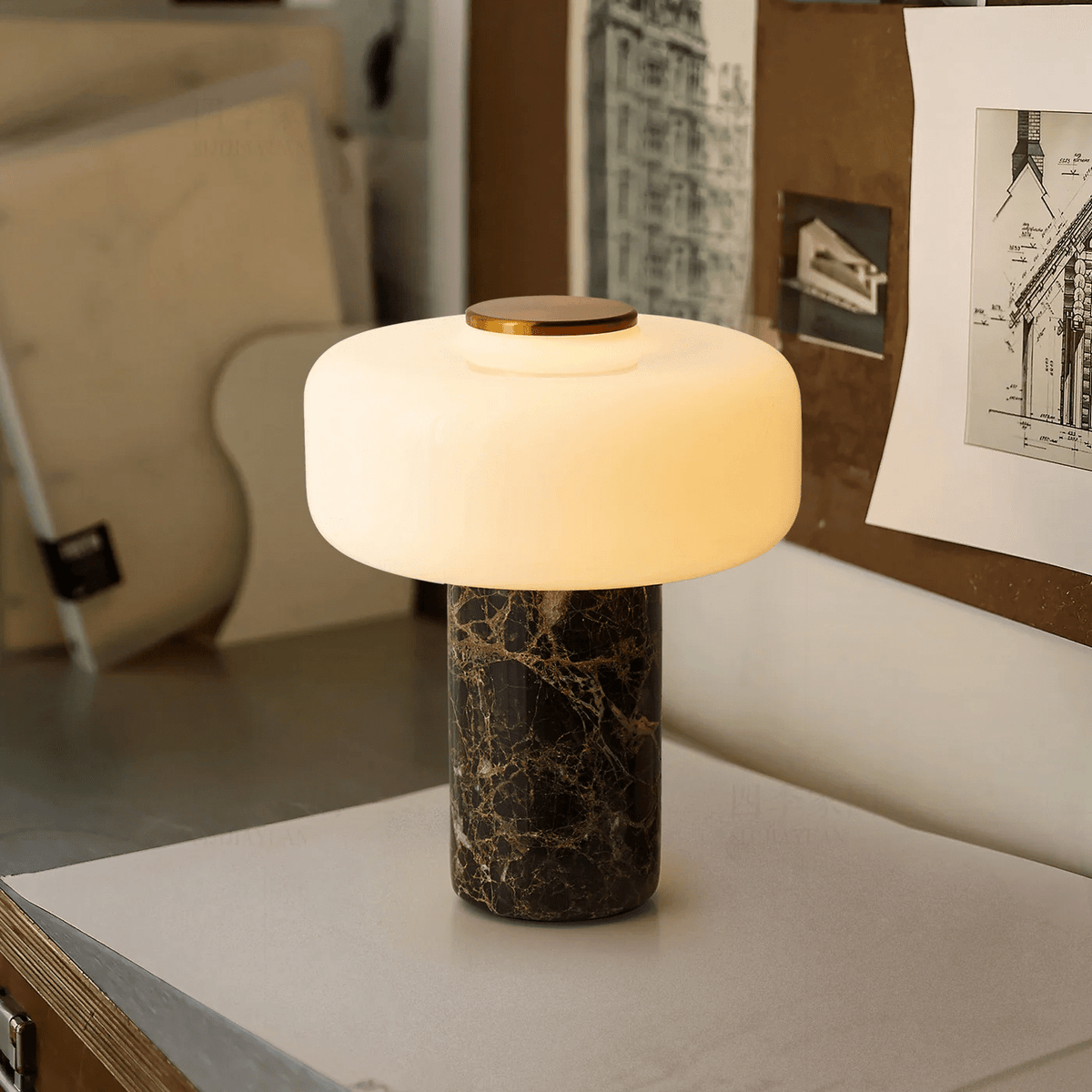A Clérachic table lamp with a dark marble base glows softly on a bench beside a wall of framed sketches.