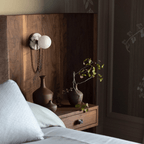 Mounted on a walnut-panelled headboard, the Clérachic sconce sits beside small ceramic vases and a folded pillow.