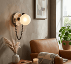 The Clérachic sconce glows on a warm plaster wall above a tan leather armchair, beside a potted plant and folded throw.