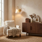 This brushed brass Clérachic wall sconce glows above a walnut sideboard beside a cream armchair draped with a throw.