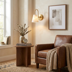 Beside a leather armchair, this brushed brass Clérachic wall sconce glows above a round side table with a small clock.