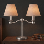 This brushed nickel Clérachic table lamp stands unlit on a dark wood table, its twin fabric drum shades visible.