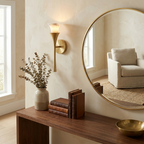 Beside a round mirror reflecting a cream armchair, this Clérachic sconce in brushed brass lights a console with books and a bowl.