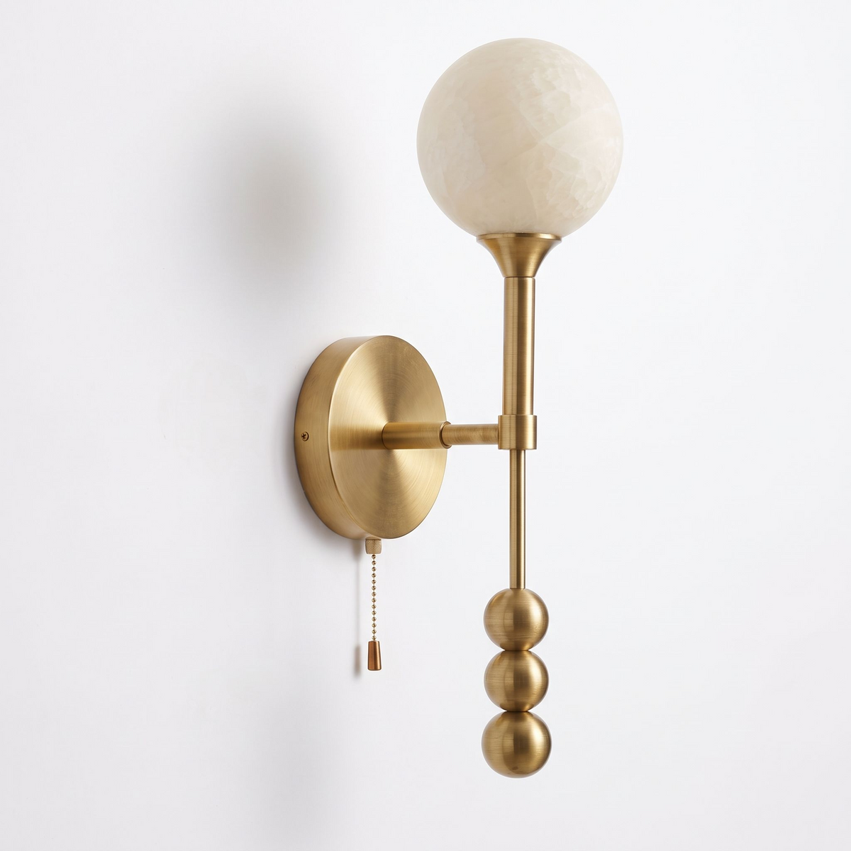 Studio shot of this Clérachic wall sconce in brushed brass, alabaster shade lit softly above the pull chain.