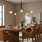A matte-black Clérachic chandelier hangs over a dining table with leather chairs and a vase of olive branches.