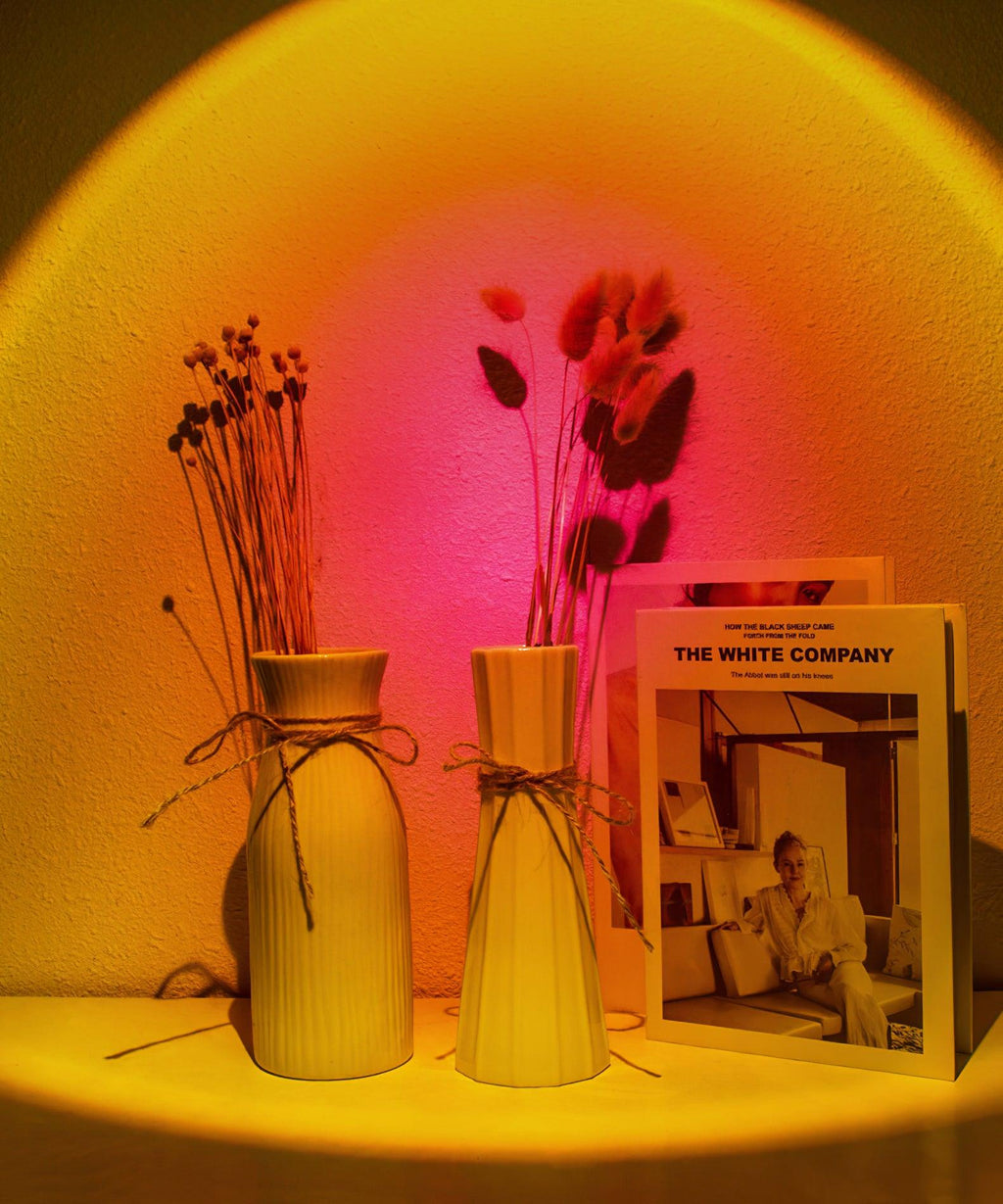 Pink and orange light from this Clérachic projection lamp fills a wall behind two vases of dried flowers and a book.