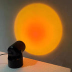 On a tabletop edge, this black Clérachic projection lamp throws a warm orange sunset halo across the wall.