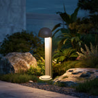 A bollard lit beside rocks and ferns at dusk, with planting lit behind it.