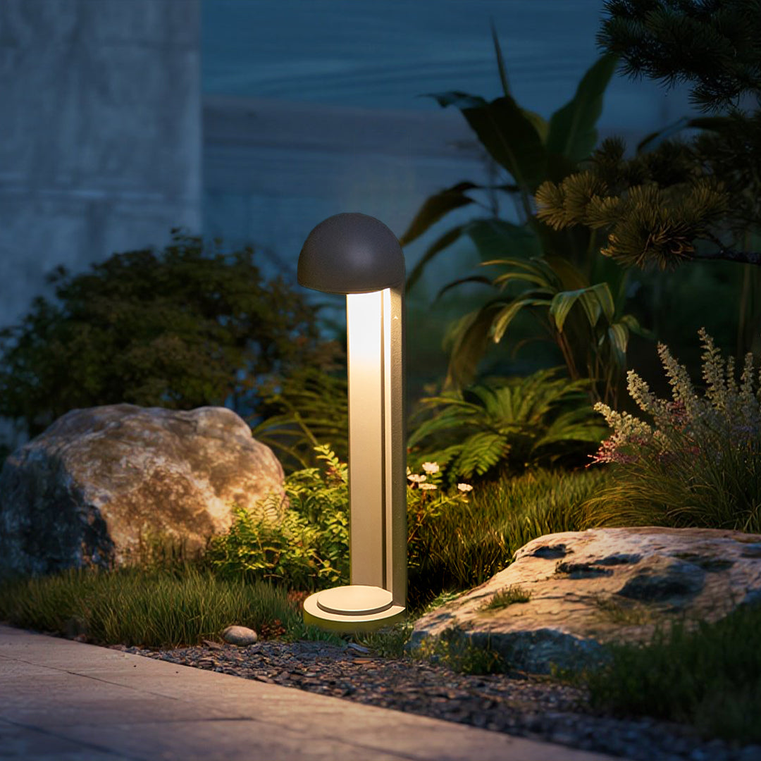 A bollard lit beside rocks and ferns at dusk, with planting lit behind it.
