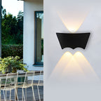 Sconce lit on a white patio wall, wire chairs and a window of the house visible to the left.