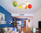 In a blue-walled living space with a bookshelf and striped rug, five Clérachic balloon lights hang in green, yellow, white, orange and red.