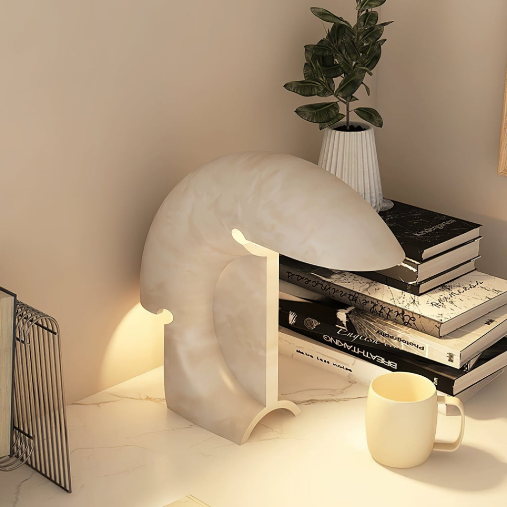 A closer view of the lit Clérachic resin lamp beside stacked books and a small plant, its crescent curve catching the warm light.