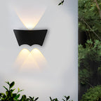 The sconce lit on a garden wall with foliage crowding in at the right, warm light fanning above and below.