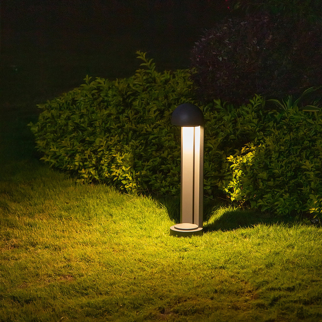 One bollard lit on grass in front of clipped shrubs, light escaping through the vertical slots.