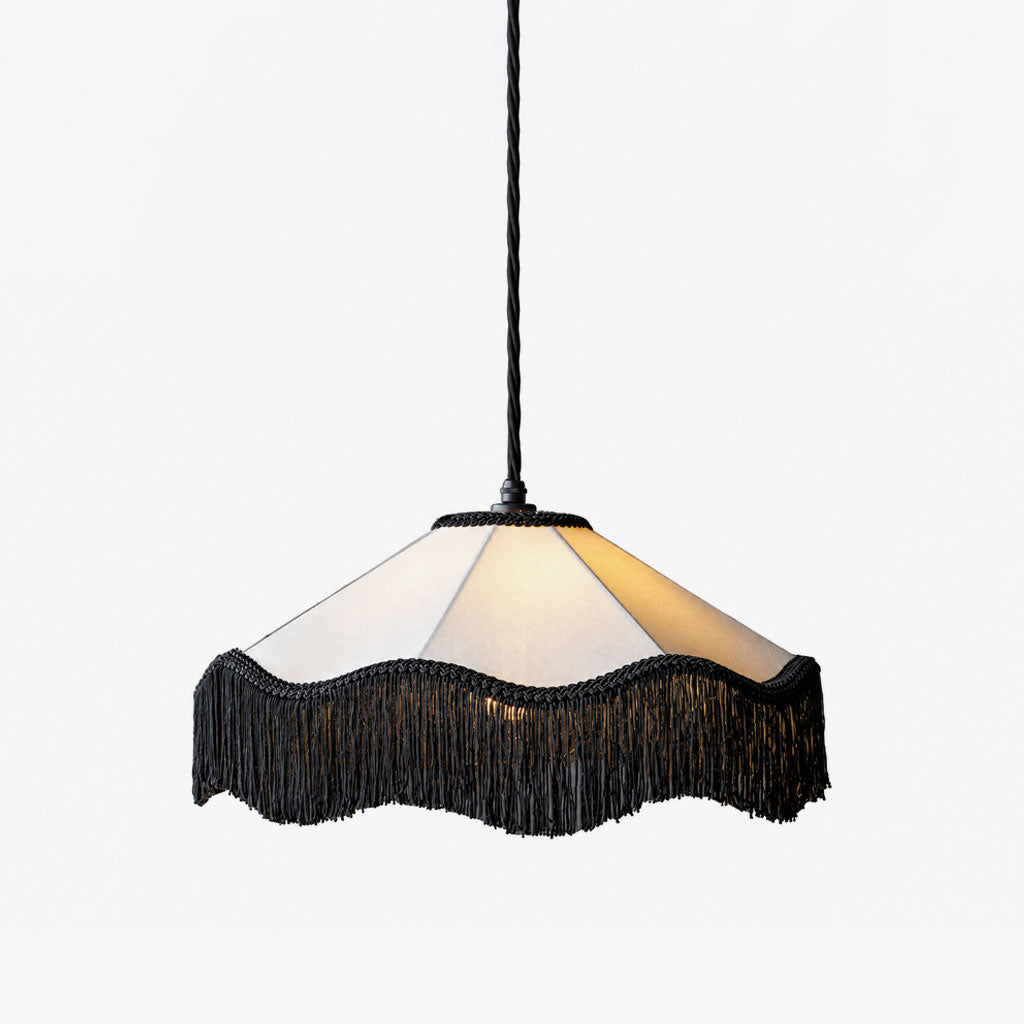 This Clérachic pendant, its faceted fabric dome edged in a black tassel fringe, shown glowing against a plain background.