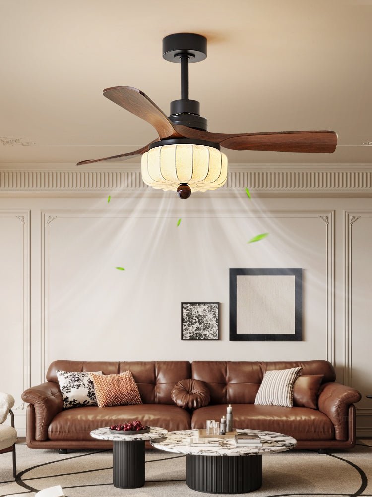 Close above a brown leather sofa with a framed print on the wall, this Clérachic ceiling fan's lantern shade glows beside its walnut blades.