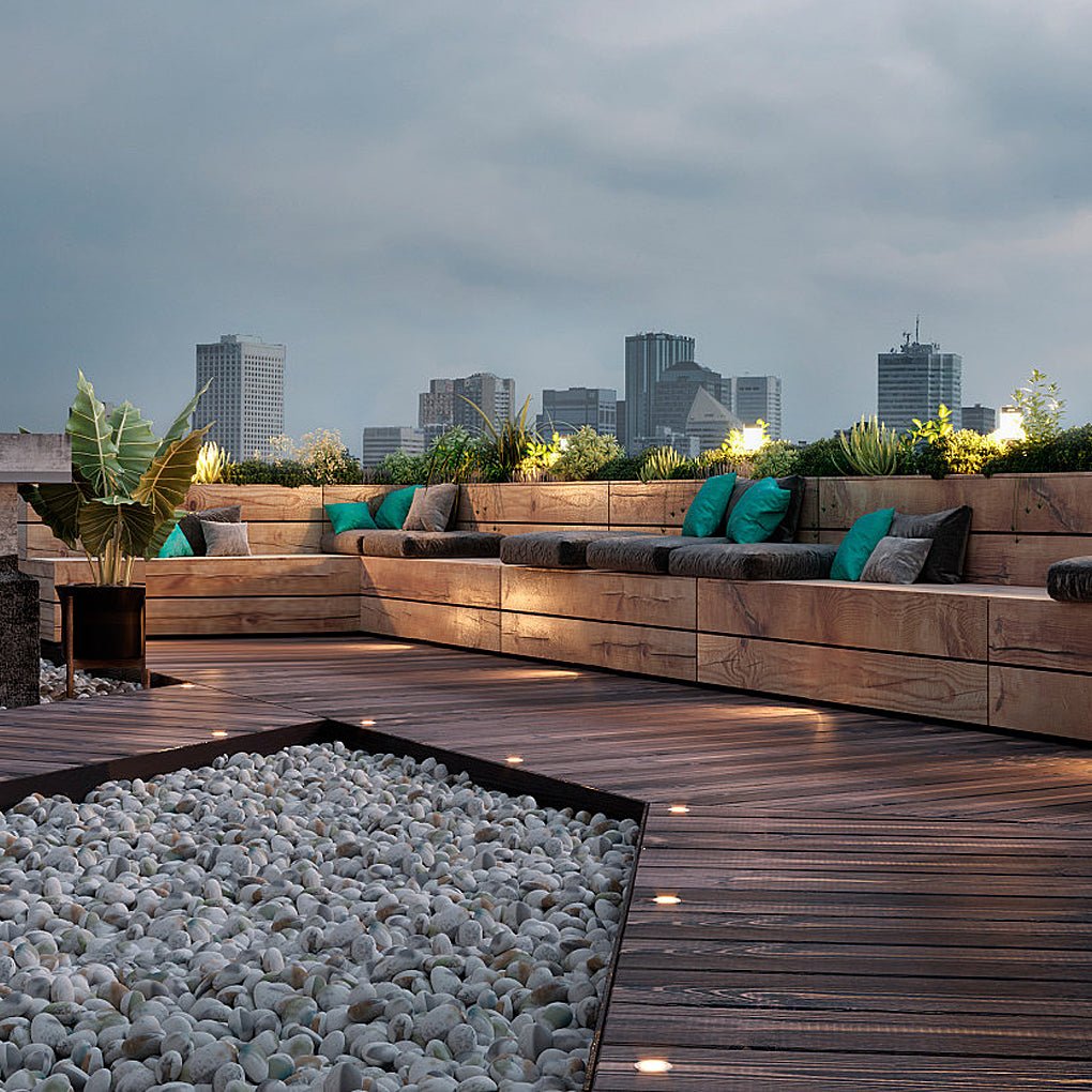 Deck lights lit into a rooftop terrace floor at dusk, with bench seating and a city skyline behind.