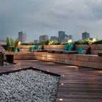 Deck lights lit into a rooftop terrace floor at dusk, with bench seating and a city skyline behind.