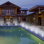 A run of lights lit along the edge of a swimming pool at dusk, in front of a timber-clad house.