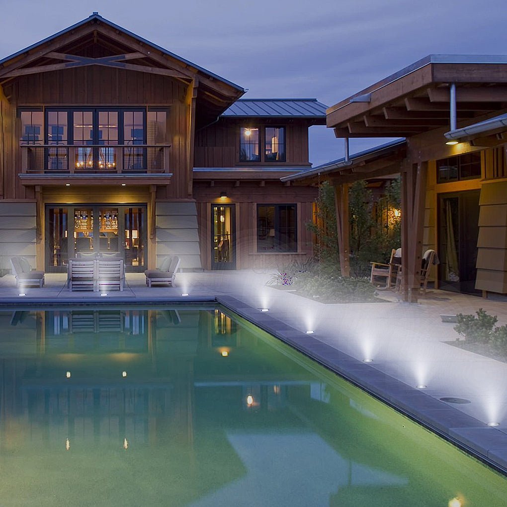A run of lights lit along the edge of a swimming pool at dusk, in front of a timber-clad house.