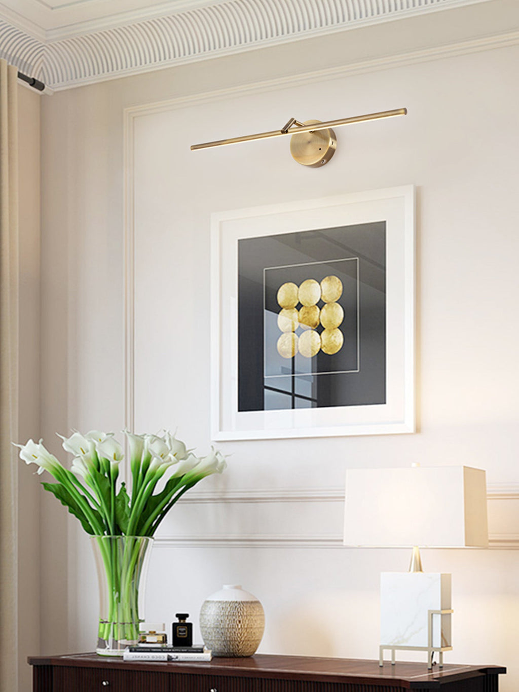 Lit gold-finish wall light from Clérachic above a framed print, with white flowers and a lamp on a dark wood console below.