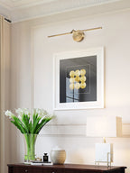 Lit gold-finish wall light from Clérachic above a framed print, with white flowers and a lamp on a dark wood console below.