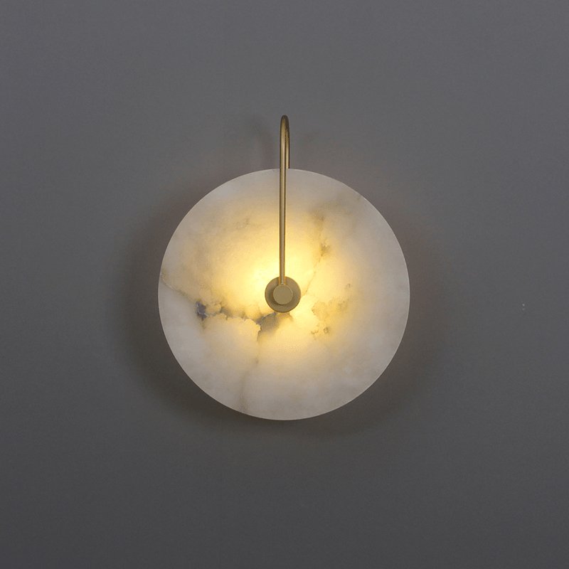 Gold-arm Elaris wall light lit against a dark grey backdrop, its darker stone veining showing through.