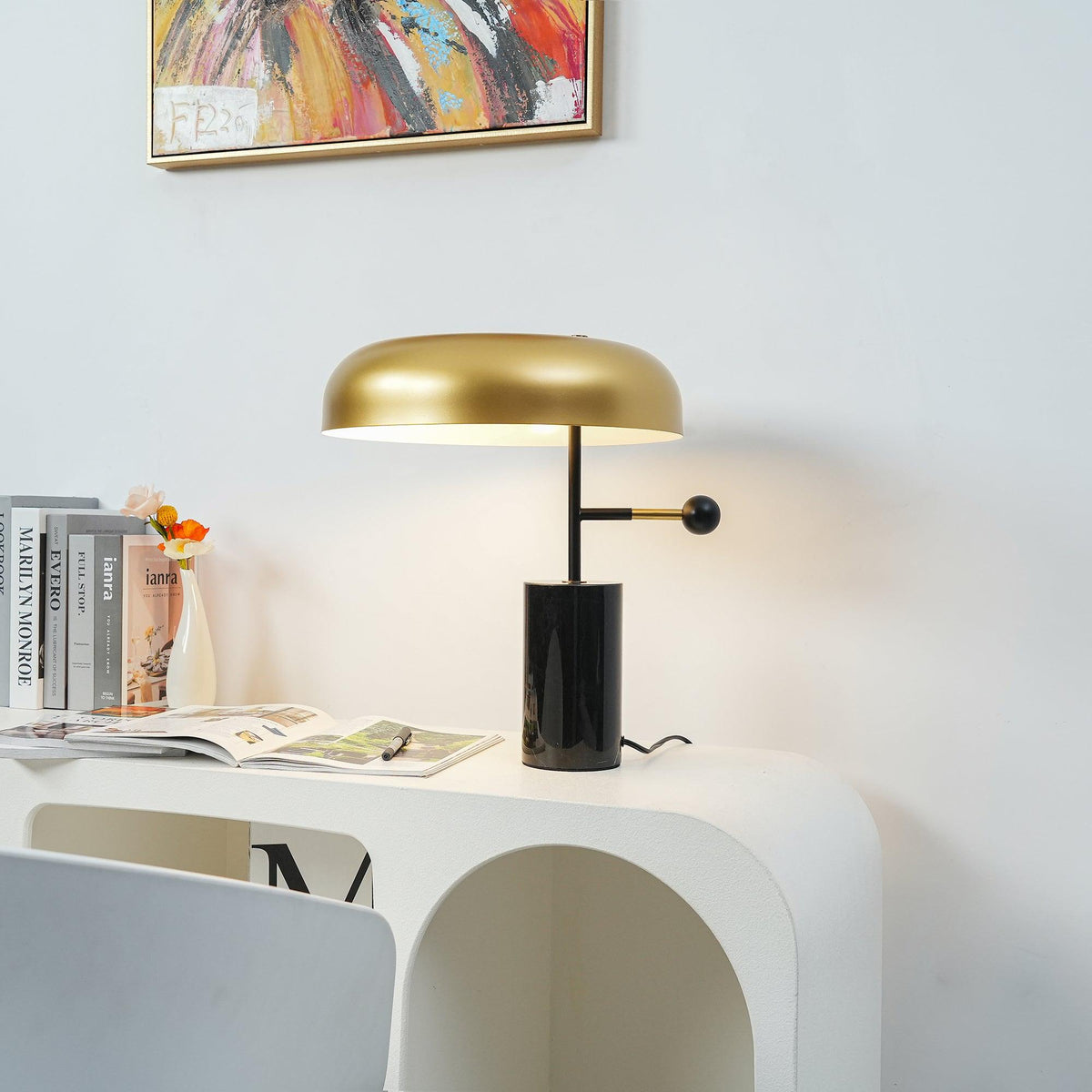 Switched on, this Clérachic table lamp casts a warm glow over books and a small vase on a curved white nightstand.