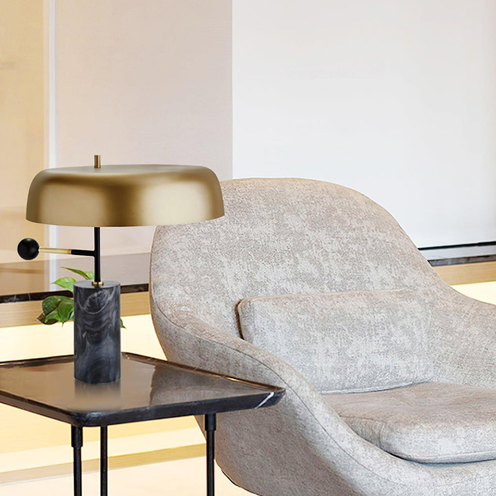 Beside a grey upholstered lounge chair, the Clérachic gold dome lamp sits on a small black side table.