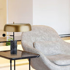Beside a grey upholstered lounge chair, the Clérachic gold dome lamp sits on a small black side table.