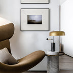 The Clérachic table lamp stands on a marble-topped side table beside a brown leather lounge chair and framed art.