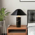 Clérachic black marble table lamp lit on a wooden nightstand beside a dark leather headboard and a wire plant stand.