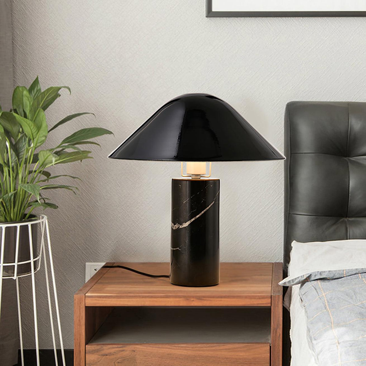 Clérachic black marble table lamp lit on a wooden nightstand beside a dark leather headboard and a wire plant stand.