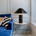 Clérachic black marble table lamp lit on a round marble side table beside a blue velvet sofa with a striped cushion.
