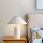 Clérachic white marble table lamp lit on a wooden nightstand beside a bed with a striped and patterned cushion.