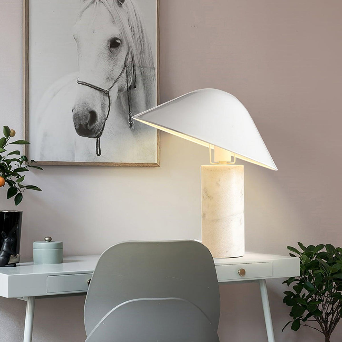 On a white desk beside a grey chair, a Clérachic white marble table lamp sits near a framed print and potted plant.
