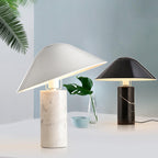 Two Clérachic table lamps, white marble in front and black marble behind, beside a monstera leaf sprig and a small blue bowl.