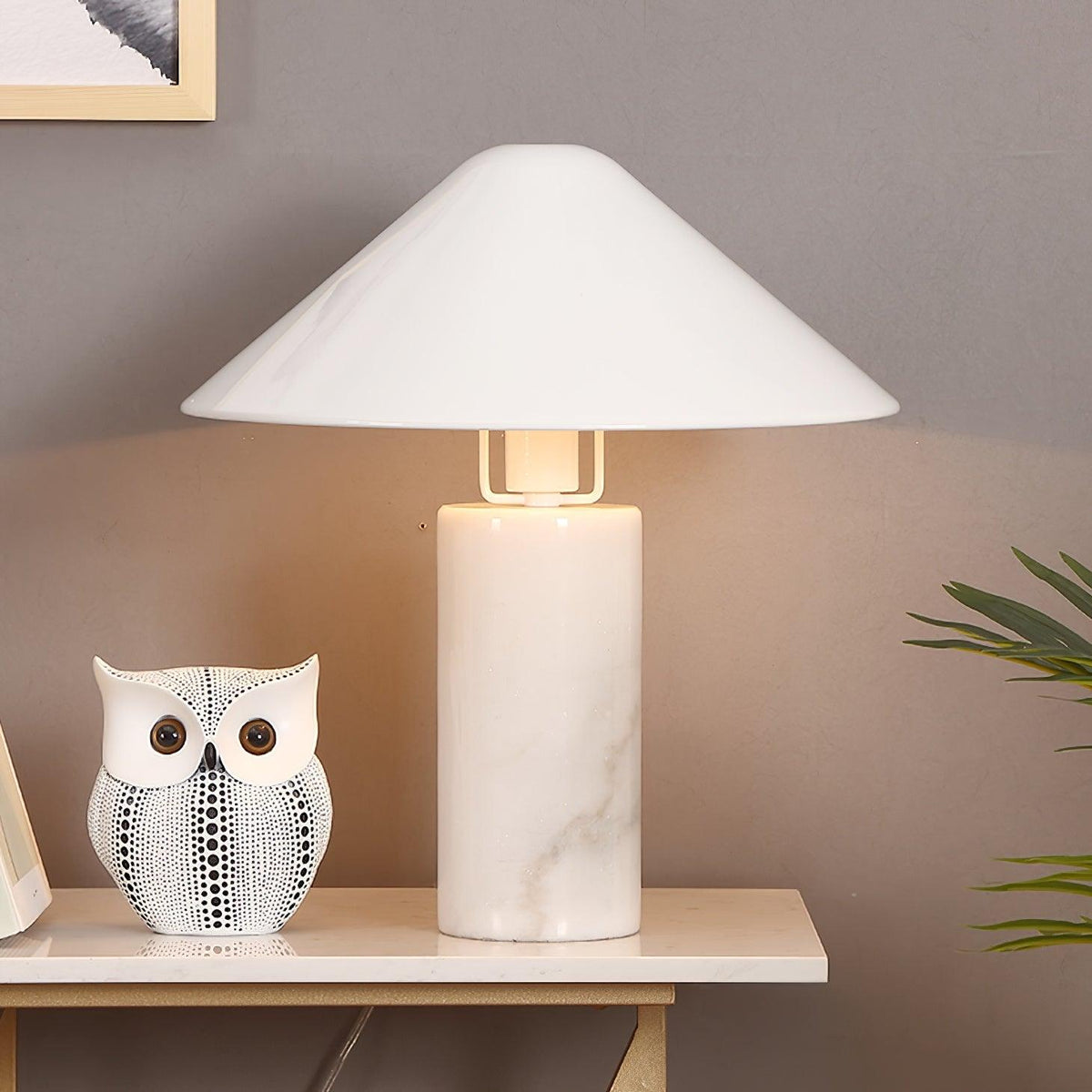 Clérachic white marble table lamp lit on a console table beside a patterned ceramic owl and trailing plant leaves.