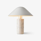 A wide angled metal shade tops a cylindrical white marble base, its warm glow visible under the rim on this Clérachic bedside table lamp.