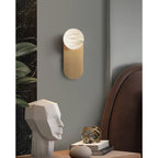 Adela alabaster wall sconce mounted on a grey bedroom wall beside a leather headboard and a tall plant